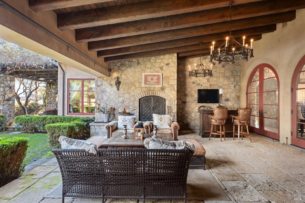 An outdoor seating area under a roof with visible beams, featuring a fireplace, cushioned seating furniture, and a bar with stools near a stone wall.