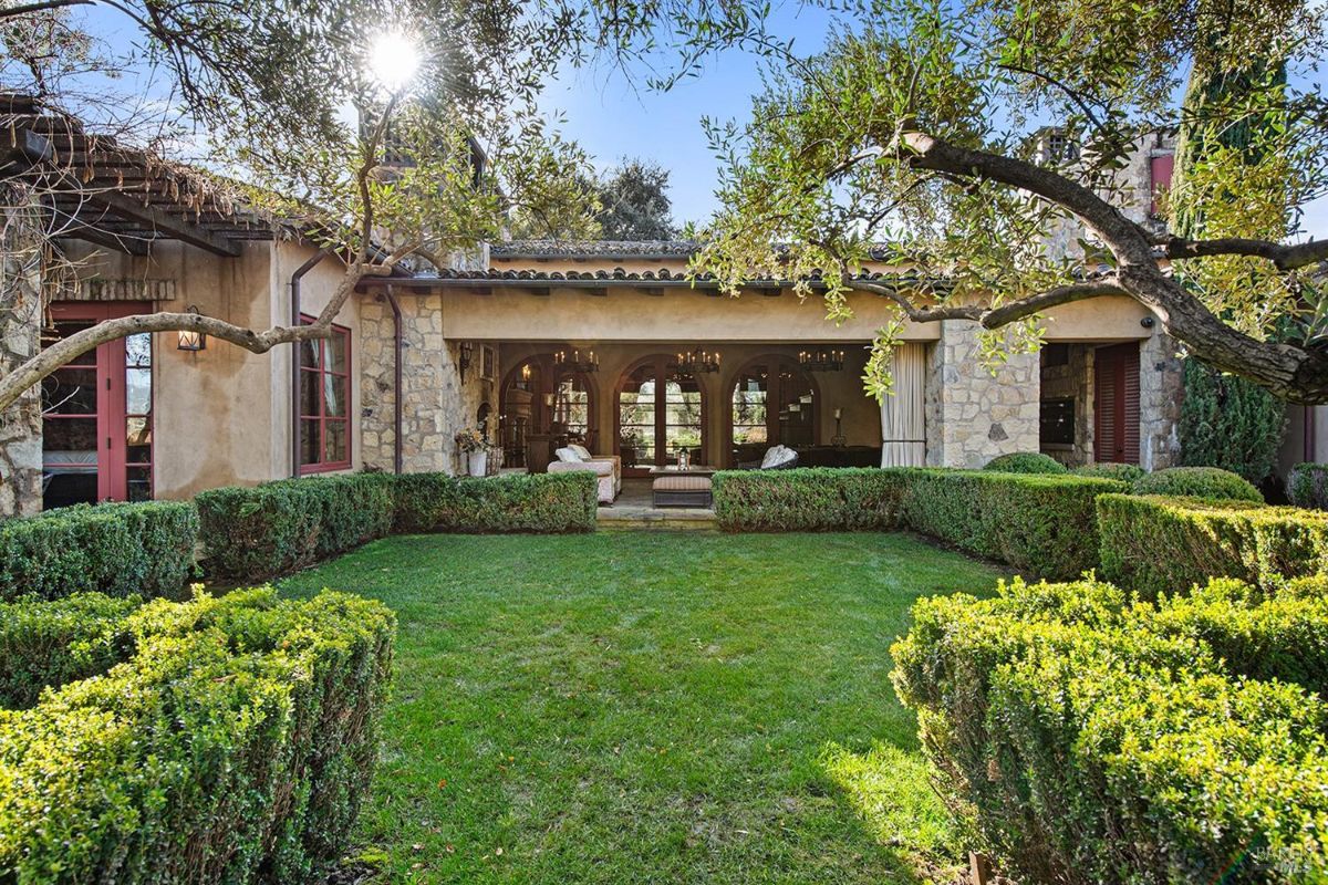 Garden features a lush, manicured lawn framed by neatly trimmed hedges. The sun shines brightly through the branches of an olive tree, casting dappled shadows on the ground. In the background, a charming stone house with arched windows and a covered patio offers a peaceful retreat. This tranquil setting evokes a sense of serenity and escape.