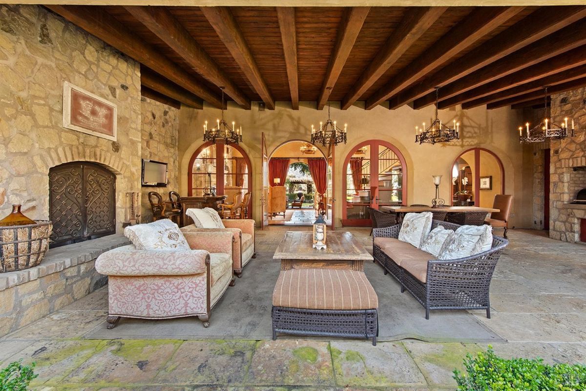 An outdoor covered seating area with stone walls, exposed wooden beams, chandeliers, and a stone fireplace. There is a mix of cushioned chairs and wicker furniture, along with a wooden coffee table.