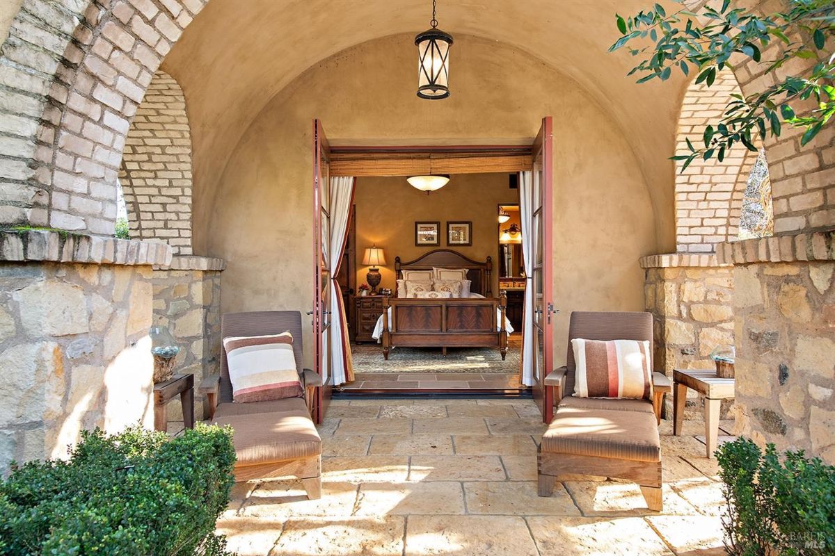 A patio area with two lounge chairs and a small side table. An arched doorway opens into a bedroom with a bed in the center.