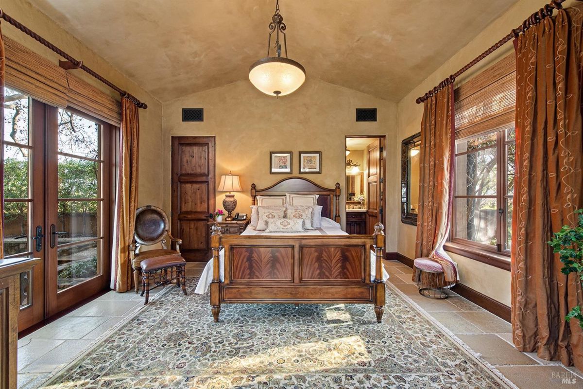 A bedroom with a bed in the center, a rug on the floor, windows with curtains, and a door leading to a bathroom.