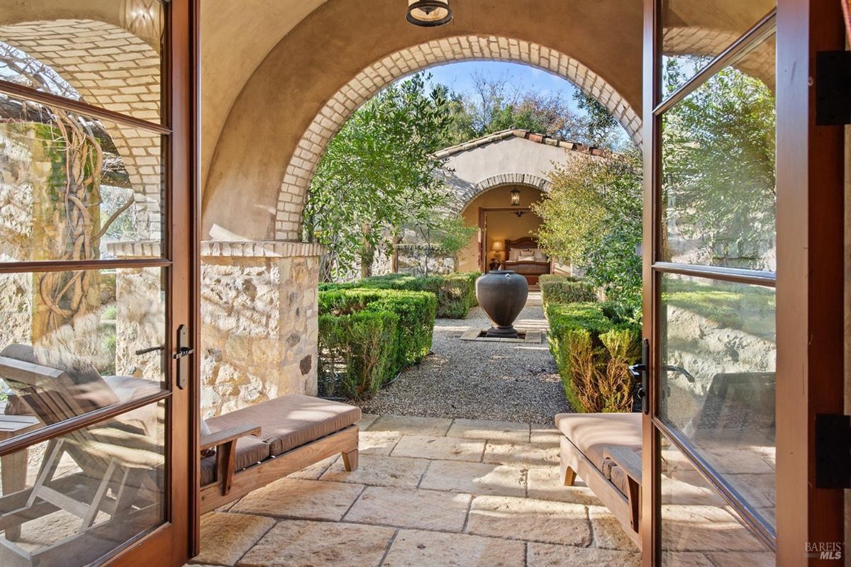 An archway view leading to a courtyard with a path lined by boxwood hedges and a central water feature. Beyond the courtyard is a room with visible furnishings.