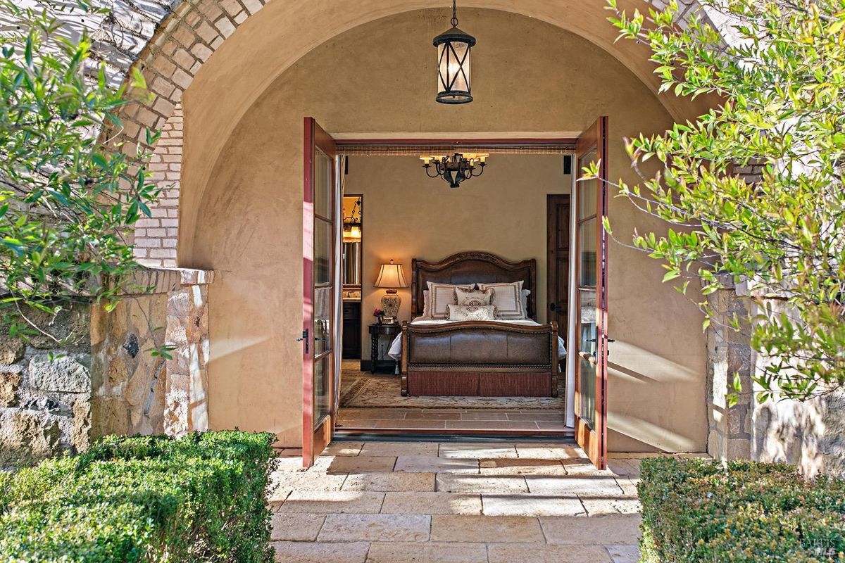 A bedroom with double doors leading outside, a bed with a leather headboard, and an arched entryway.