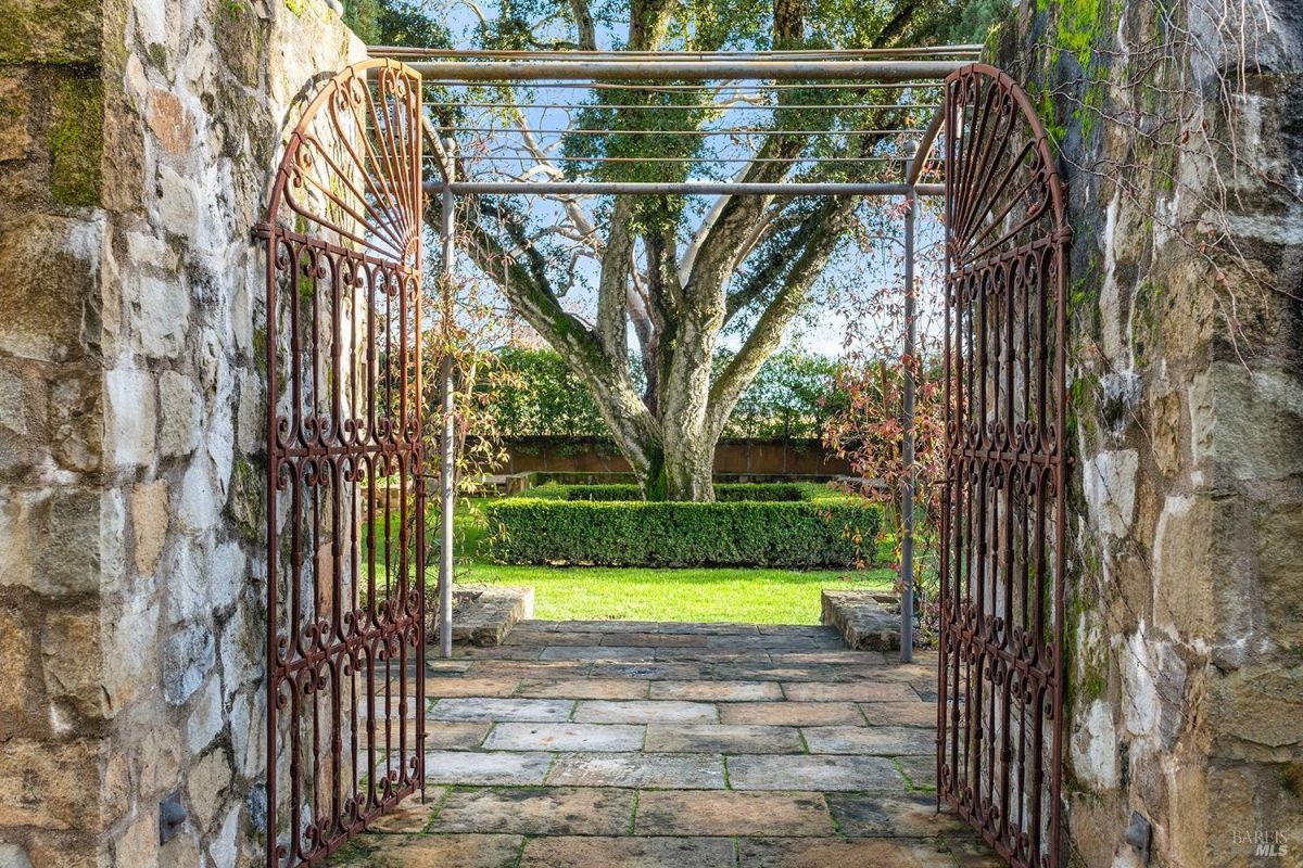 Wrought iron gates, framed by weathered stone walls, open to reveal a secret garden, inviting exploration and promising tranquility. The ancient tree at its heart, with sunlight dappling the manicured grounds, beckons visitors to discover the hidden beauty within.