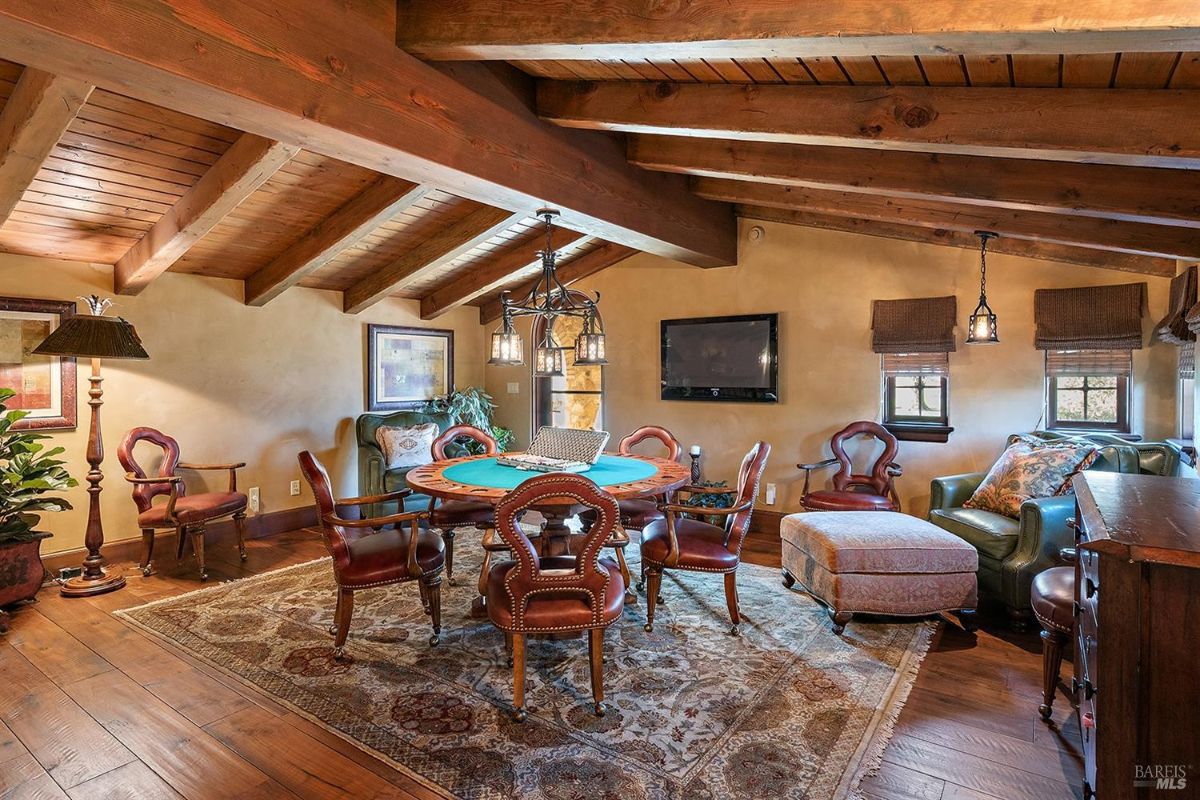 A smaller room with a wooden ceiling and beams. The space includes a table with chairs, upholstered seating, and a rug. Windows and wall-mounted light fixtures are visible.