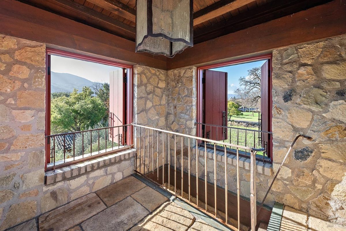 An interior space with stone walls and two open windows with red wooden shutters. A metal railing runs along the edge of the windows. The floor is made of stone tiles, and there is a hanging light fixture on the ceiling. The windows provide views of outdoor greenery and distant hills.