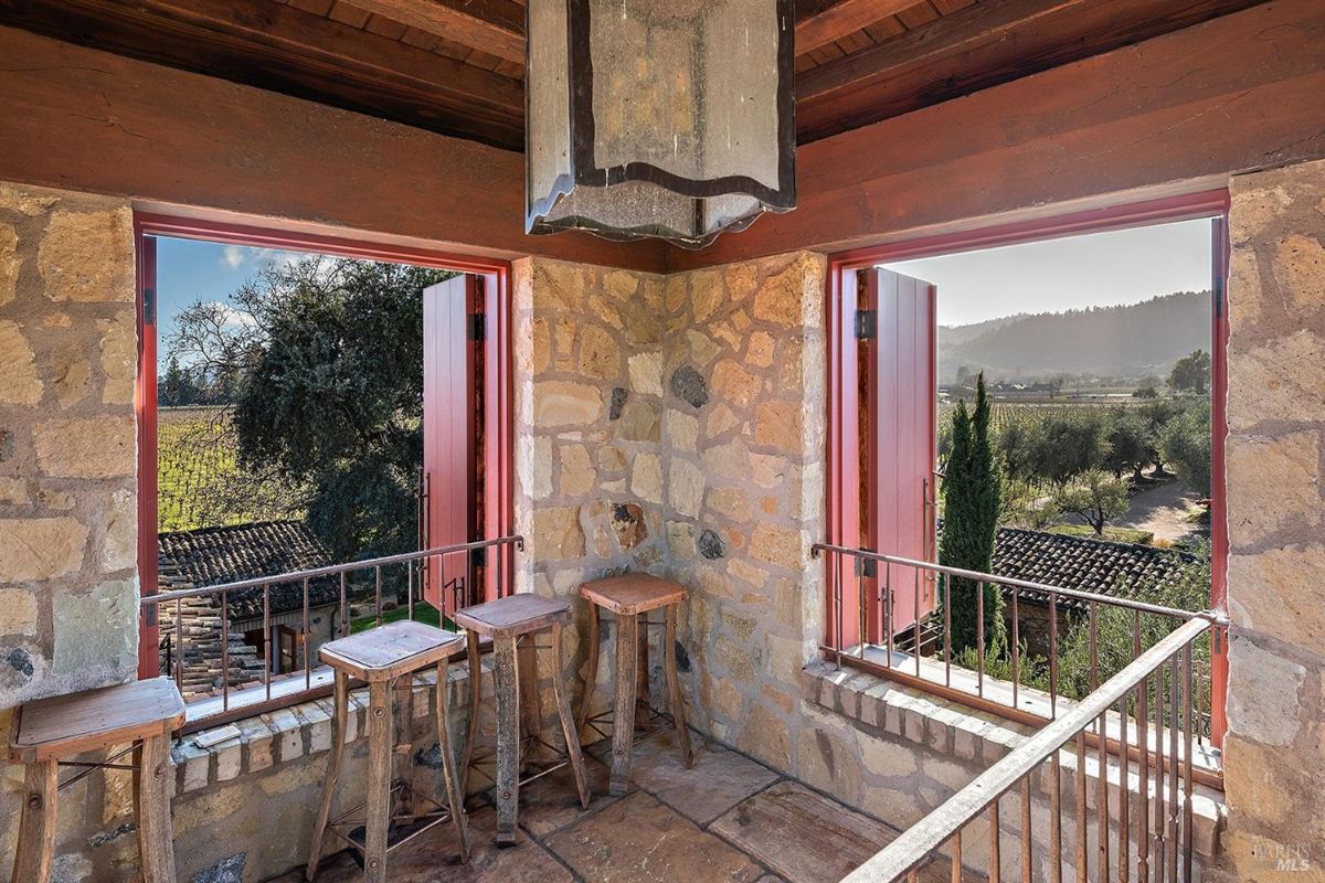 An upper-level interior space with stone walls, wooden beams, and small wooden stools. The space has large open windows overlooking the surroundings.