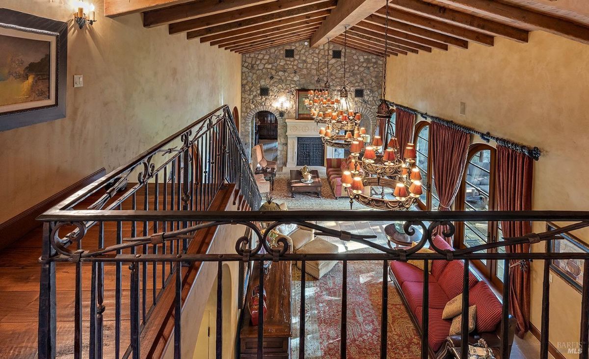  A view of a living room from an upstairs railing showing chandeliers, a stone fireplace, and large arched windows with curtains.