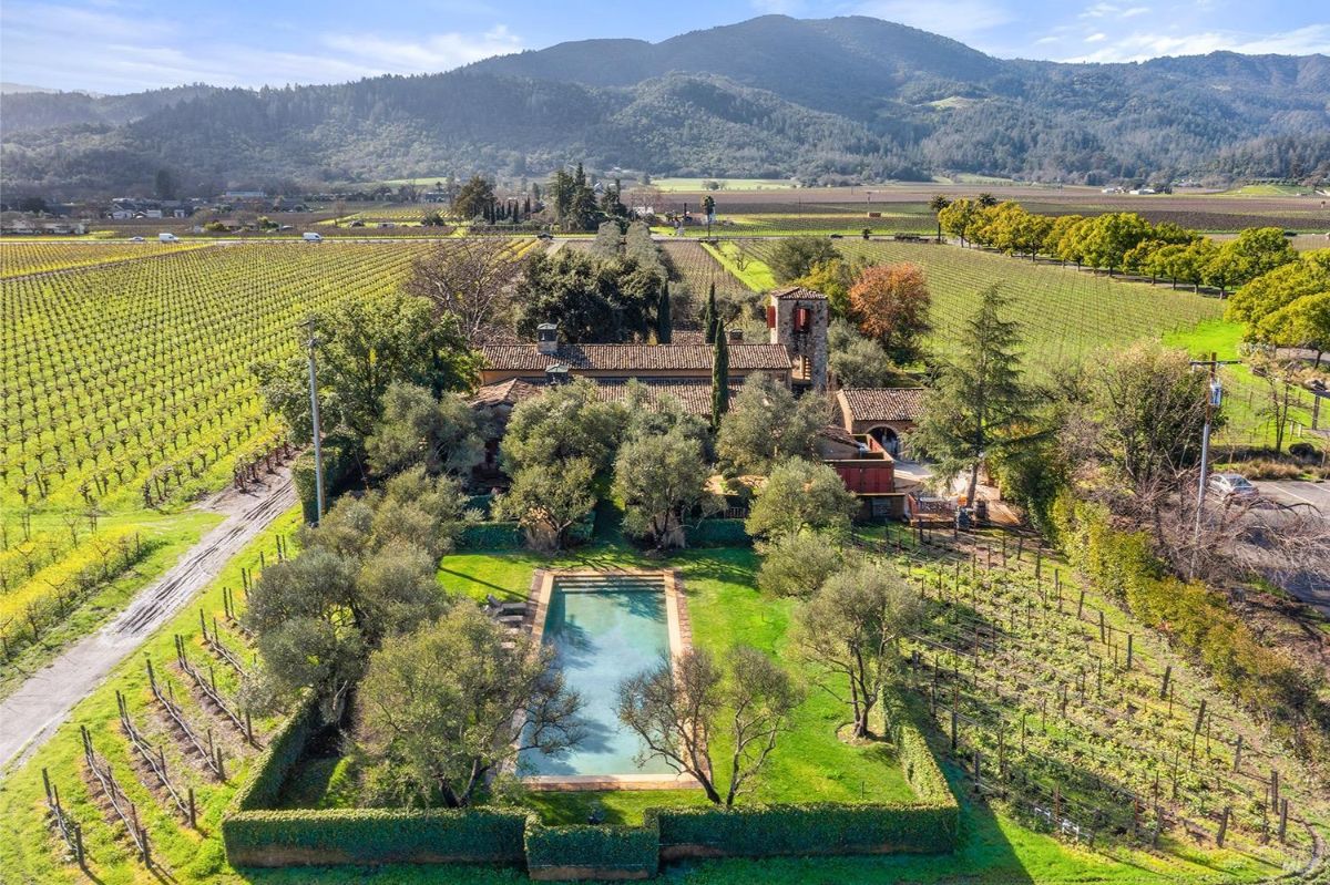 Aerial view of a property surrounded by vineyards and fields. There is a rectangular pool in the foreground, bordered by greenery, and a large stone structure with tile roofing in the background.