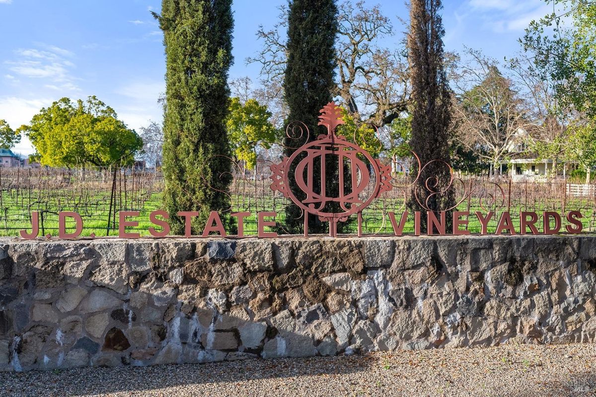 A stately stone wall, bearing the elegant wrought iron insignia and name of "J.D. Estate Vineyards," marks the entrance to this distinguished winery. The vineyard stretches out behind, hinting at the meticulous craftsmanship and dedication to producing fine wines.