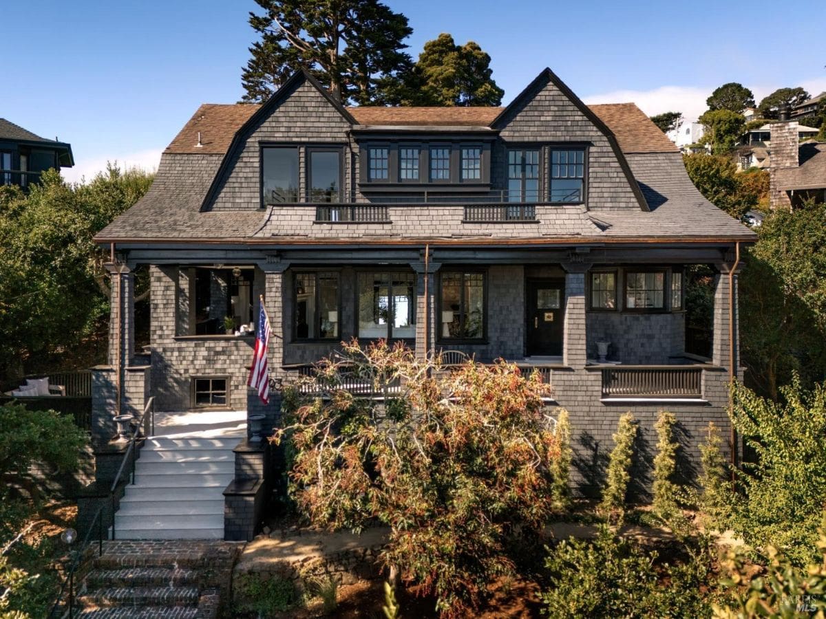 A gray shingle-style house with a wide front porch and surrounding trees.