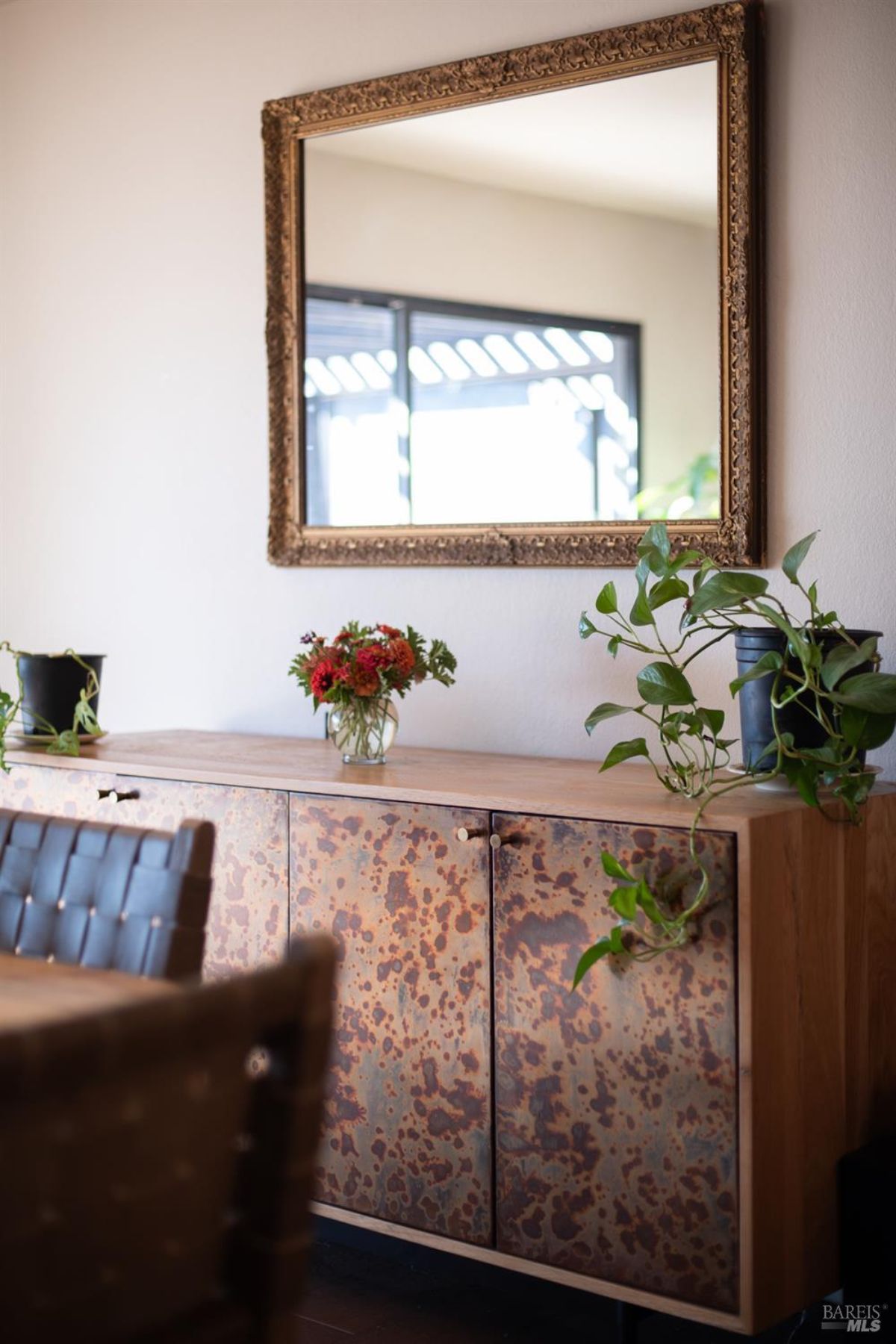 Sideboard with metallic-textured doors and plants, topped with a mirror and flower arrangement.