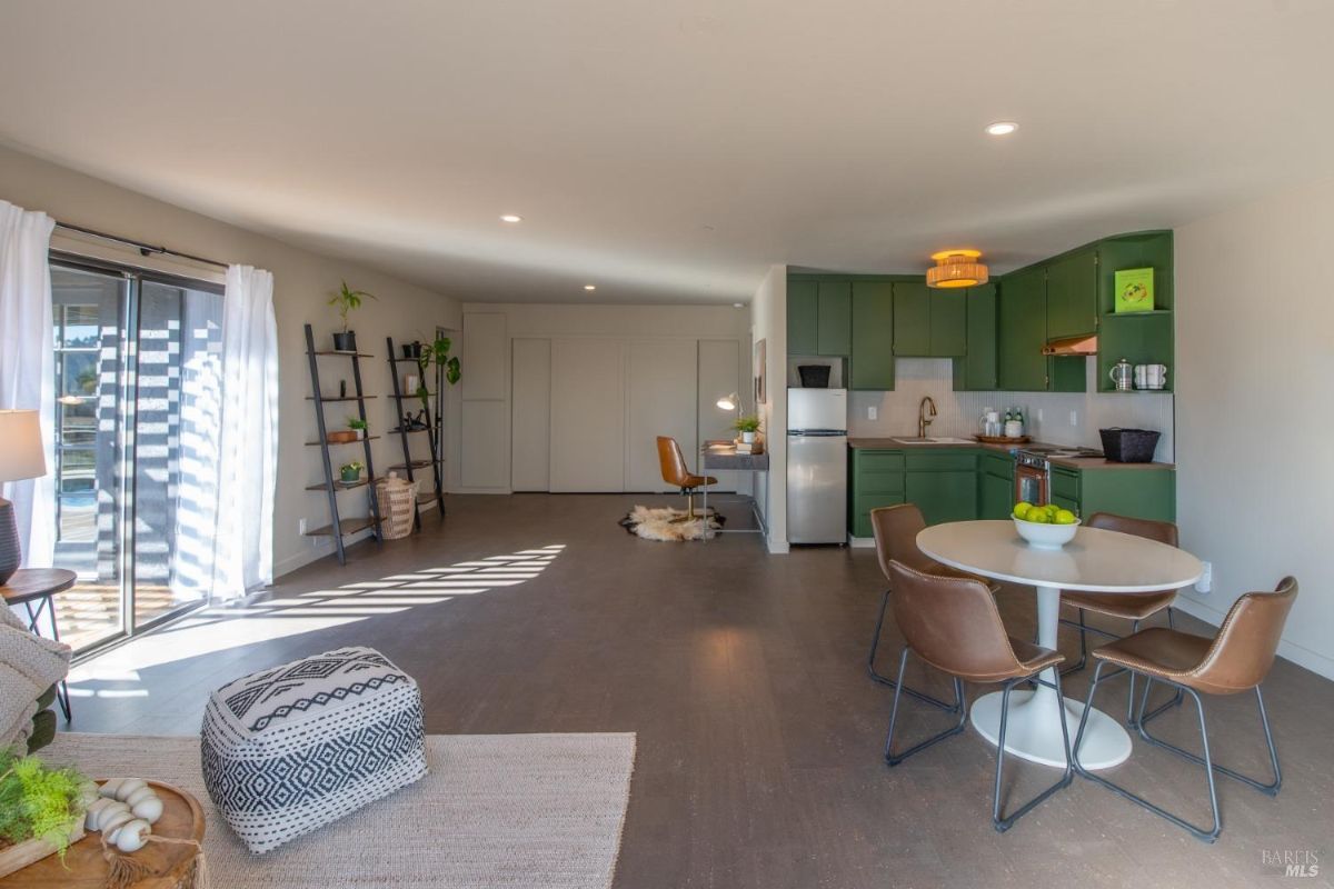 Open kitchen and living area with green cabinetry, dining table, and a seating corner, with natural light streaming in.