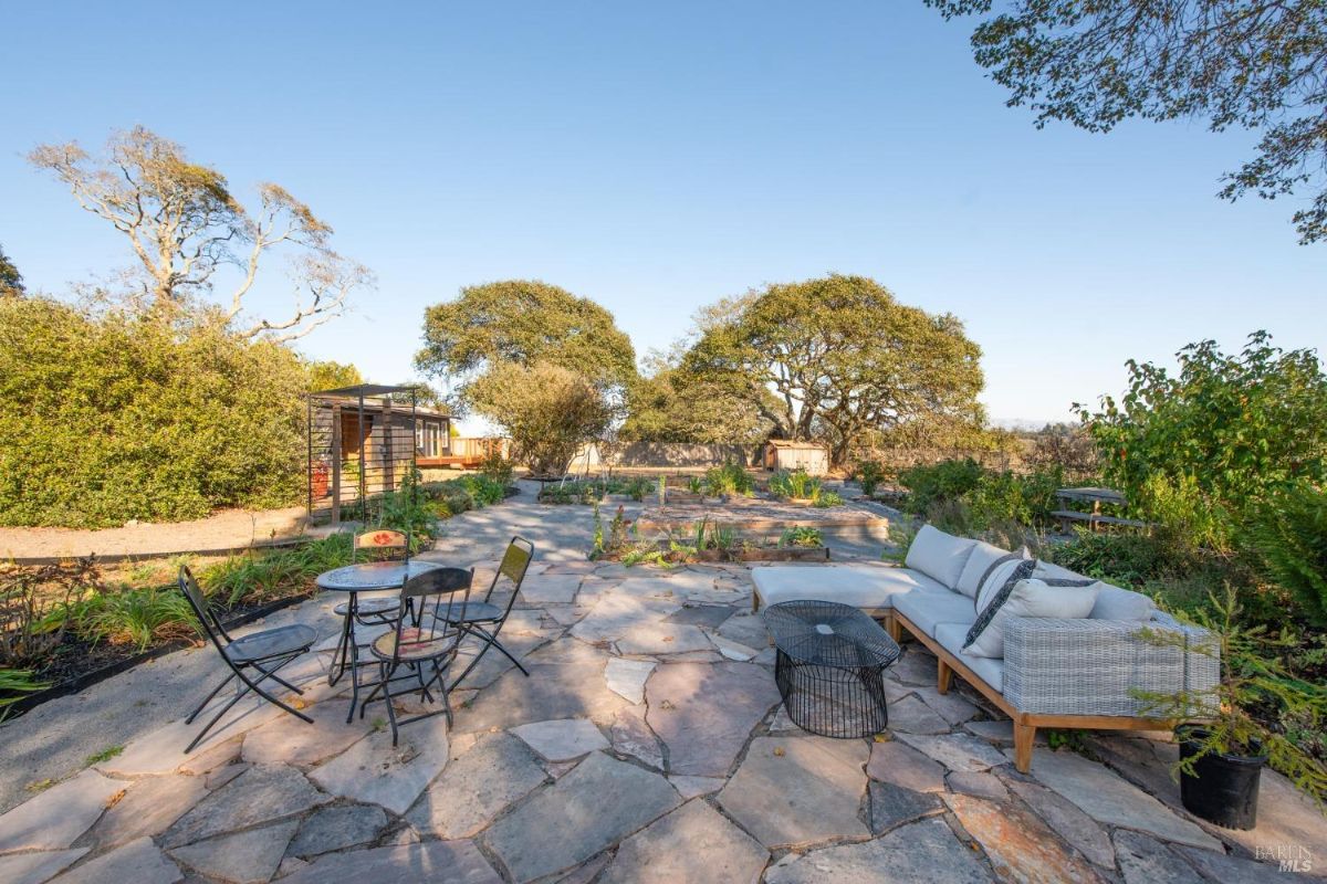 Stone patio area with seating, surrounded by gardens and trees.