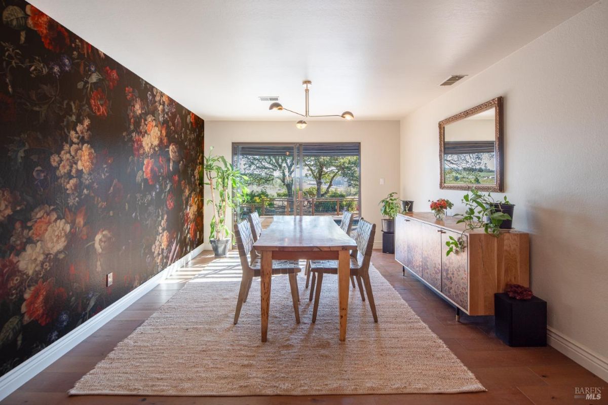 Long dining table with wooden chairs set against a wall featuring floral wallpaper and large sliding glass doors.