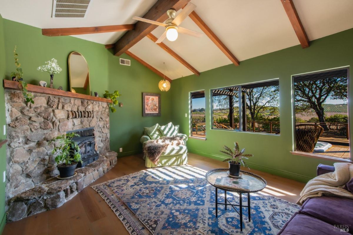 Living room with green walls, wooden ceiling beams, a stone fireplace, and large windows overlooking the outdoors.