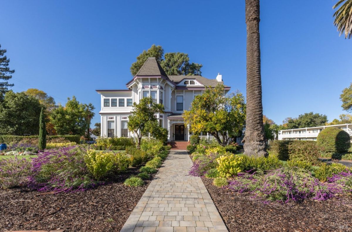  a closer view of the entrance with double doors and a porch. Brick steps and pillars are visible along with surrounding vegetation. The front garden contains fruit trees and flowering plants.