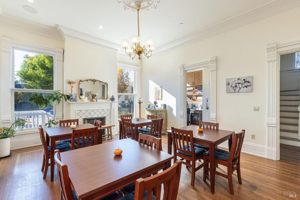 This room contains multiple small wooden dining tables with matching chairs, arranged for seating. A decorative fireplace with a mantel and mirror is centered on one wall, flanked by large windows allowing natural light. An open doorway leads to a kitchen, and a staircase is visible through another open passageway.