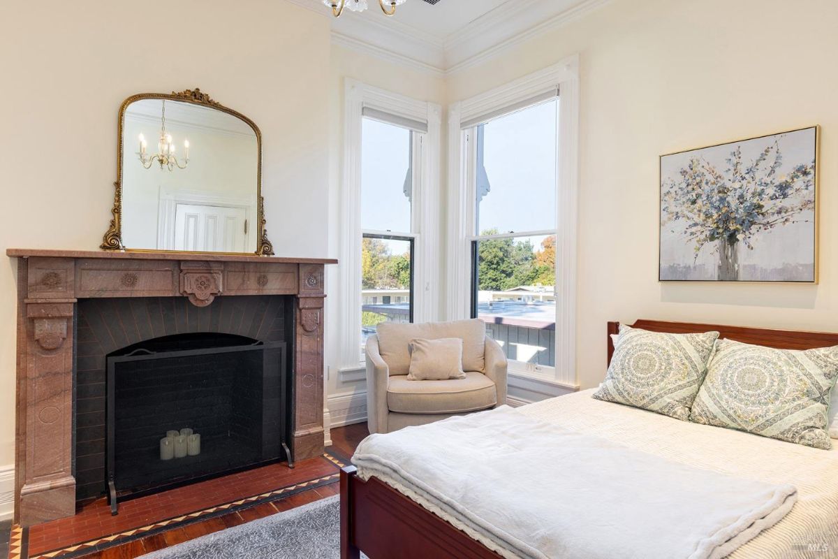 A cozy bedroom featuring a wooden bed, a soft armchair near large windows, and a vintage fireplace adorned with a golden mirror. The room feels bright and airy with natural light pouring in. 