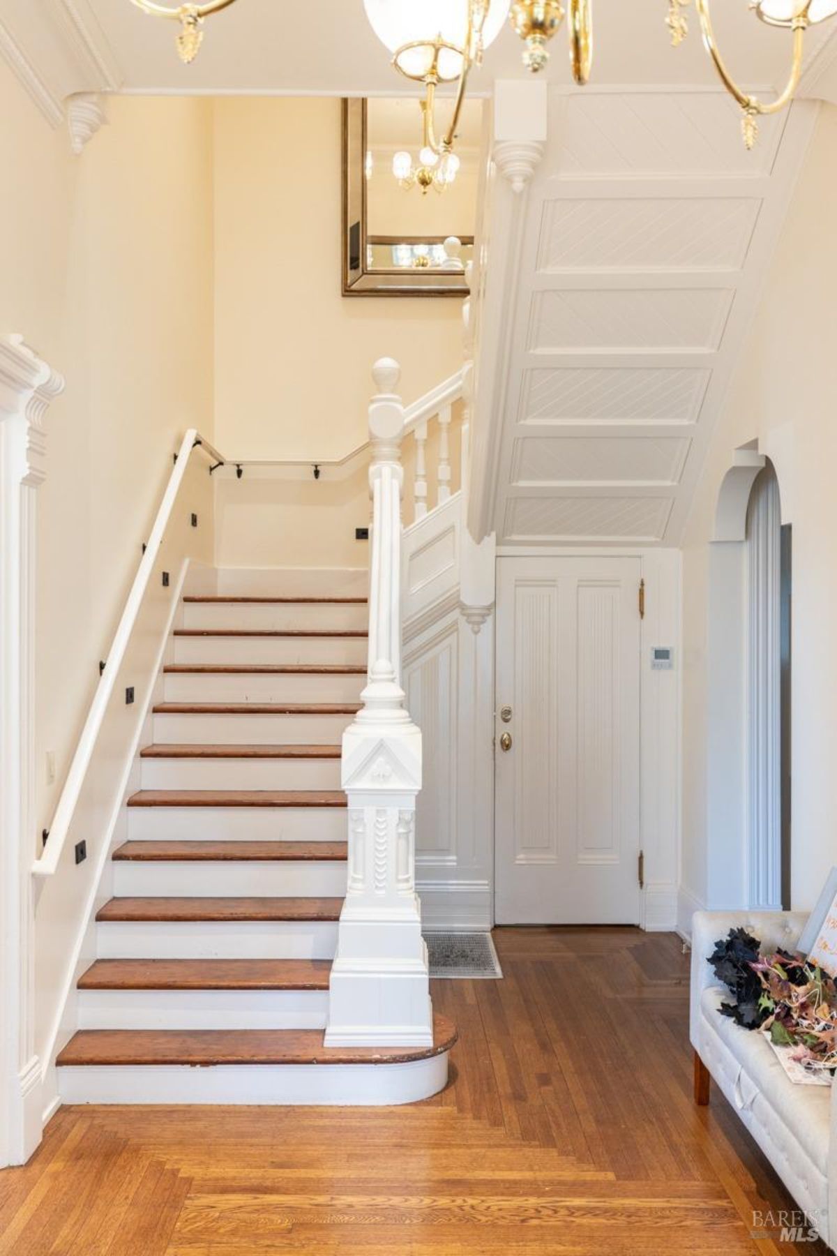 Another view of the staircase shows the surrounding entryway, with a chandelier hanging from the ceiling and an elegant coffered design overhead. The space is warm and inviting with its natural wood floors.