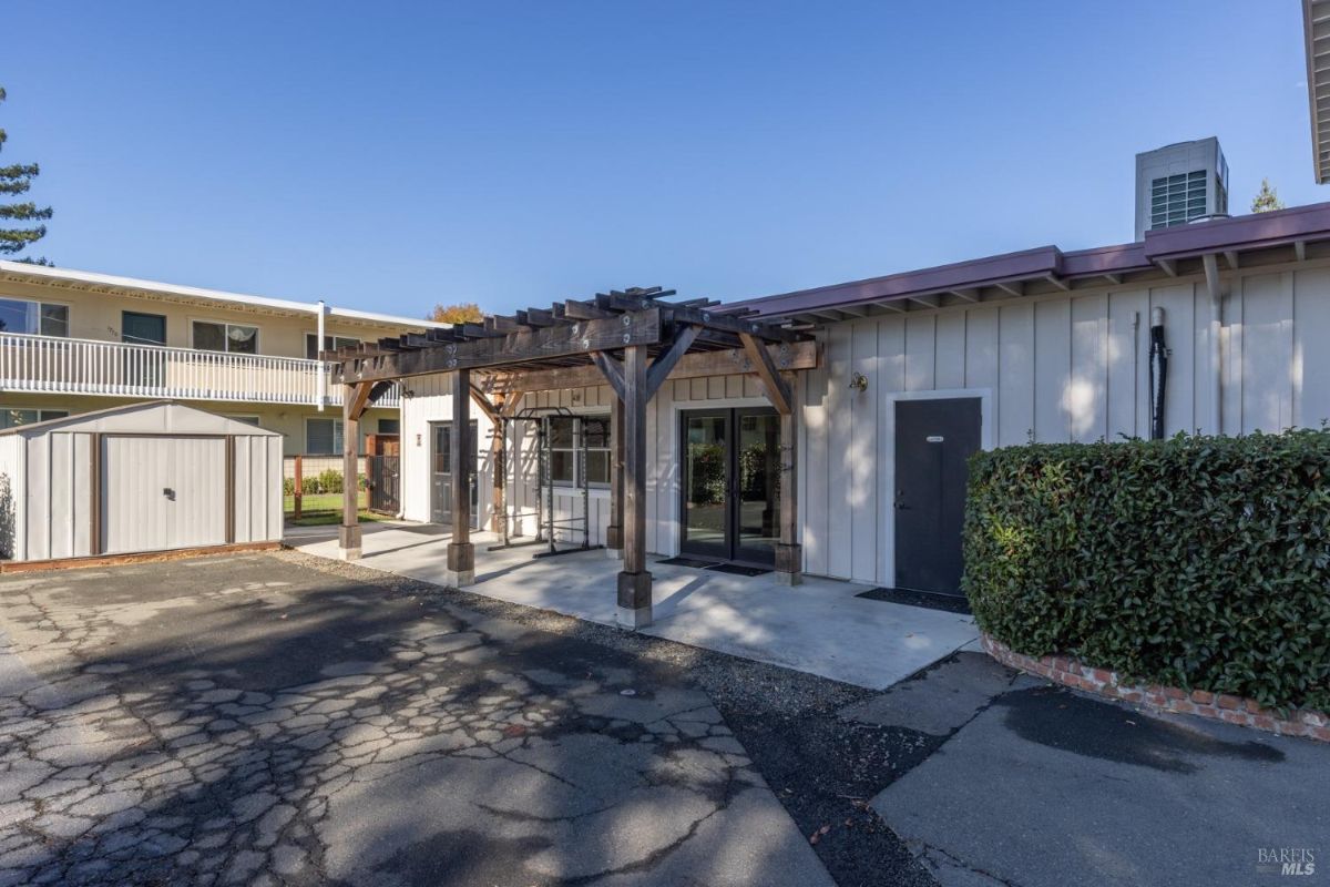 A pergola attached to a modern structure, likely serving as an outdoor seating or functional area. The space includes a storage shed, paved ground, and trimmed hedges, blending utility and aesthetics. The surrounding buildings appear to be part of a residential or mixed-use complex. The pergola adds a rustic touch to the otherwise clean-lined design.