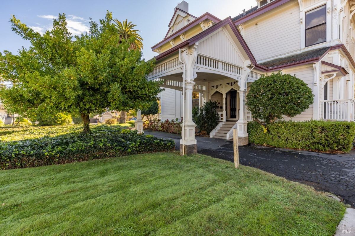  This image showcases the front entrance of a classic Victorian-style house. The structure features ornate wooden detailing, a covered porch, and well-maintained landscaping with a lush lawn and mature trees. The light exterior paint contrasts beautifully with the greenery, giving the home a charming and inviting appearance.