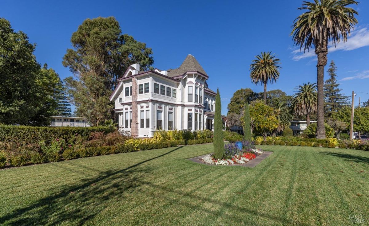  The image highlights the front garden and entrance of the house. A paved pathway leads to the front porch, with shrubs, trees, and flower beds lining the approach. The architecture includes decorative trim and large windows.