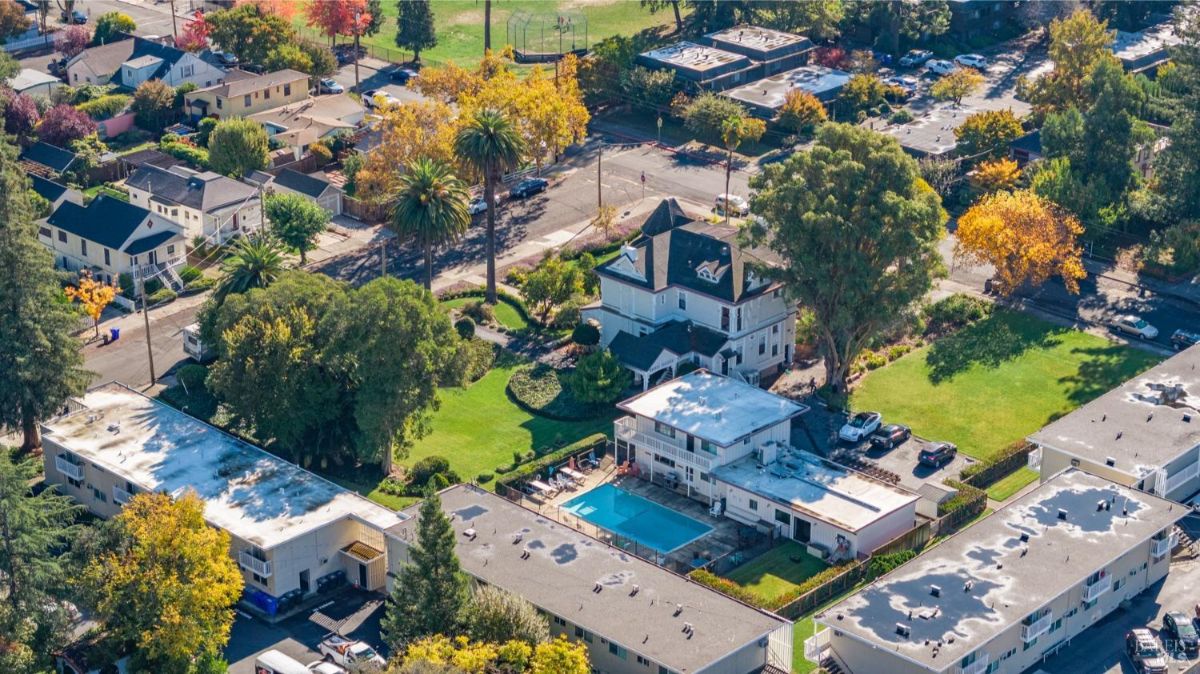  Aerial view displays the Victorian-style house within its neighborhood. The property includes a pool area and landscaped grounds, and is situated among multi-family residential units. Streets and surrounding community features are also visible.