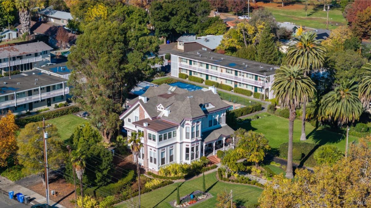 This image captures the house from an elevated angle, focusing on its detailed exterior features. It shows the landscaped grounds with palm trees and shrubbery surrounding the house. Nearby residential buildings and open spaces are visible in the background.