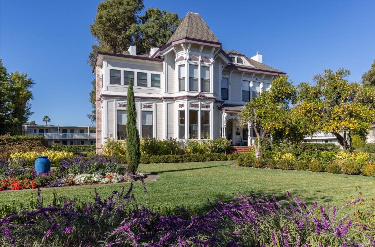 A closer view of the entrance with double doors and a porch. Brick steps and pillars are visible along with surrounding vegetation. The front garden contains fruit trees and flowering plants.