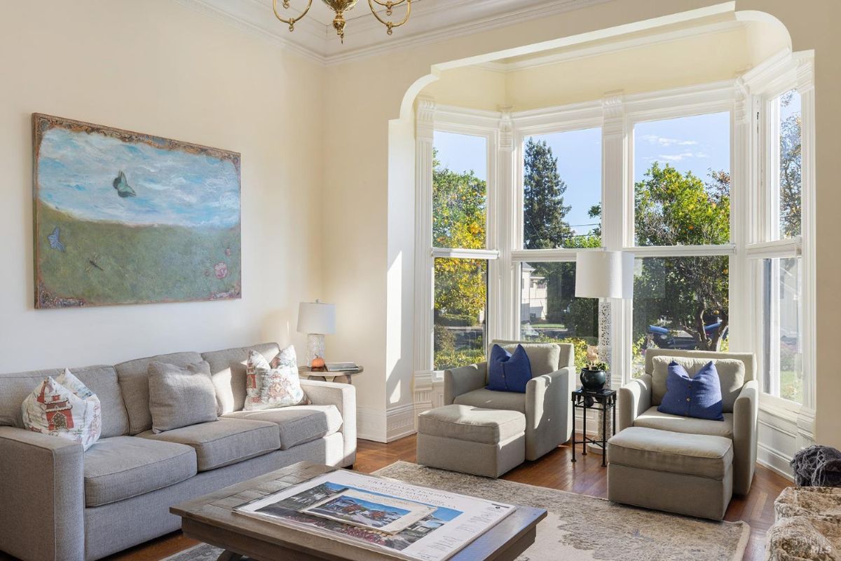  A different angle of the living room shows a seating arrangement positioned near a large window. The window overlooks the garden outside, and the room is brightly lit. A table is in the center, with a painting displayed on the wall.