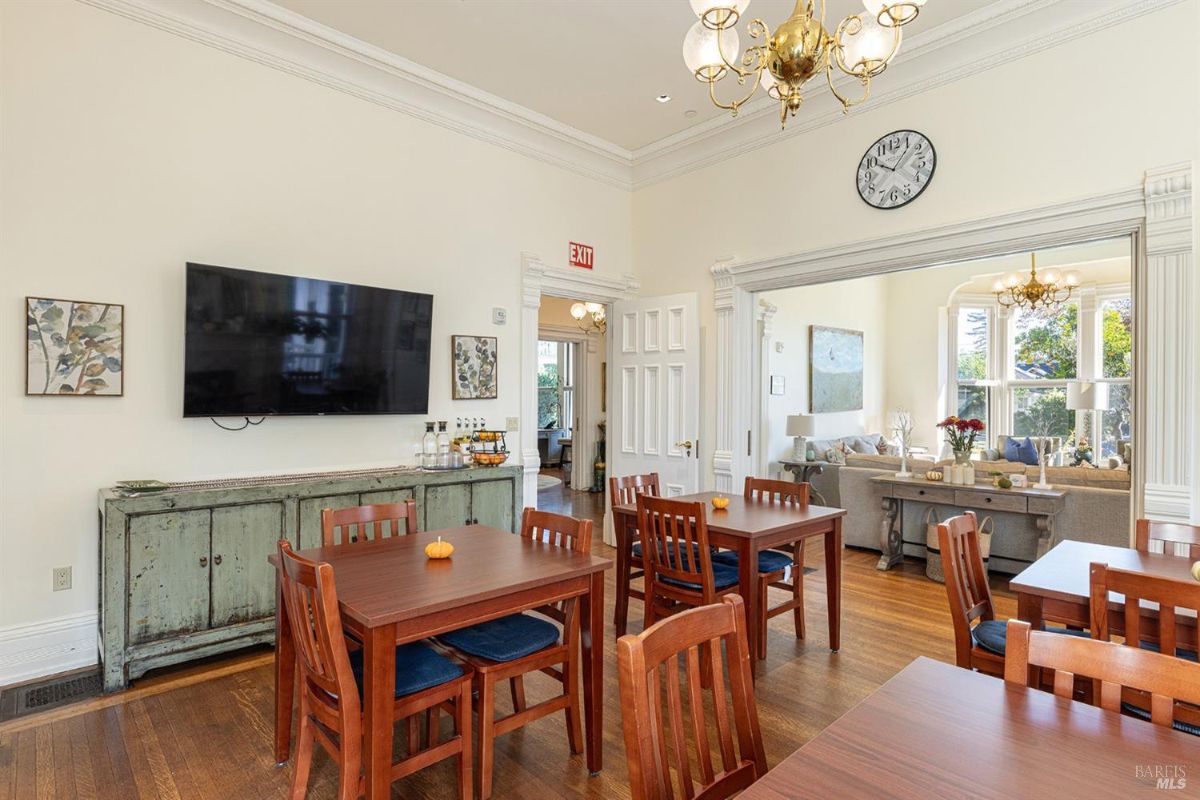 This space features several wooden dining tables with chairs, arranged in a compact format. A large television is mounted on the wall above a weathered cabinet, with a clock placed above it. A wide opening with decorative trim provides a view into a sitting area with sofas and large windows.