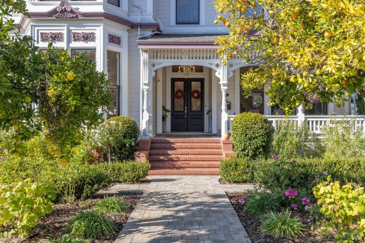 a closer view of the entrance with double doors and a porch. Brick steps and pillars are visible along with surrounding vegetation. The front garden contains fruit trees and flowering plants.