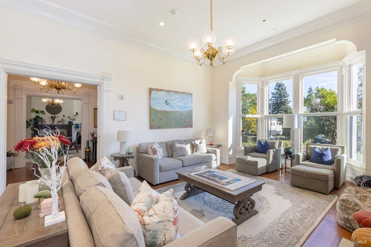 The interior of the house shows a living room with seating arrangements, a large bay window, and a chandelier. Light streams through the windows, illuminating the space. Artwork and decorative elements are present on the walls and furniture.