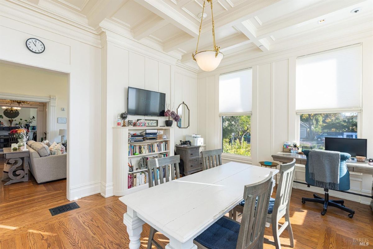 A cozier room with a rectangular table surrounded by chairs. Shelves hold books and decor, while windows offer a serene view of the outdoors. The white walls and ceiling moldings create an elegant, clean look.