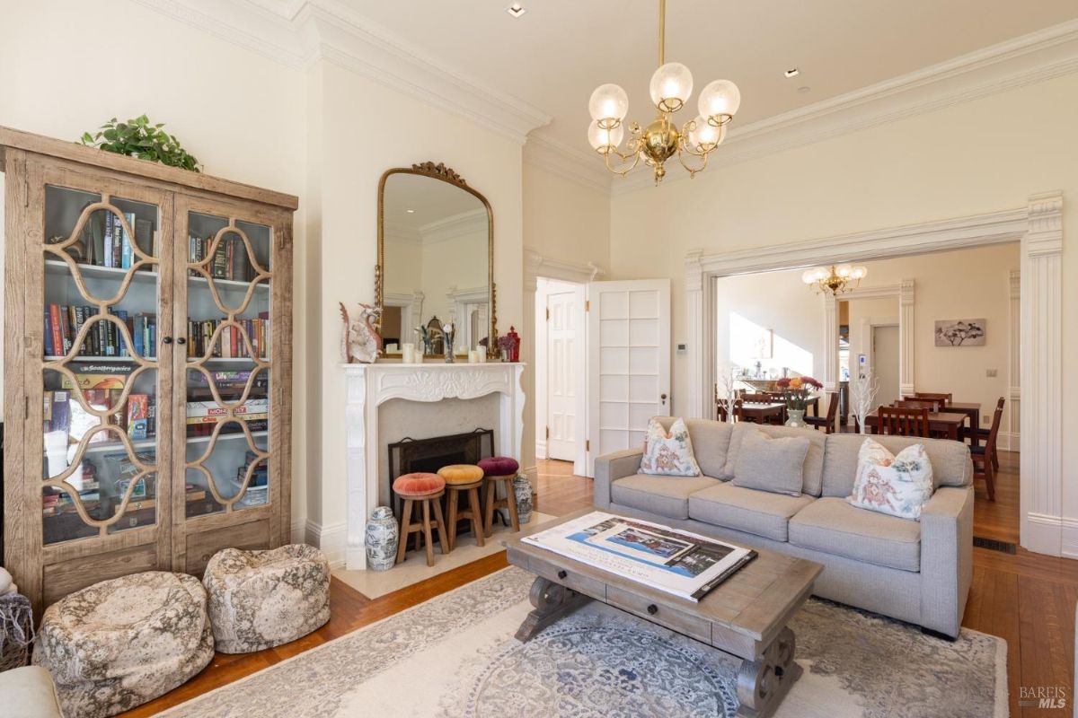 This image shows another section of the interior, featuring a fireplace, seating area, and bookshelf. Doorways lead to adjacent rooms, and a chandelier is suspended from the ceiling. The flooring and trim are consistent with traditional designs.