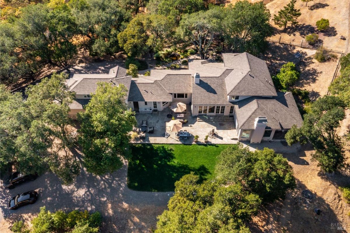 A large residential property with a gray roof, surrounded by trees, and a backyard with a lawn and patio area.