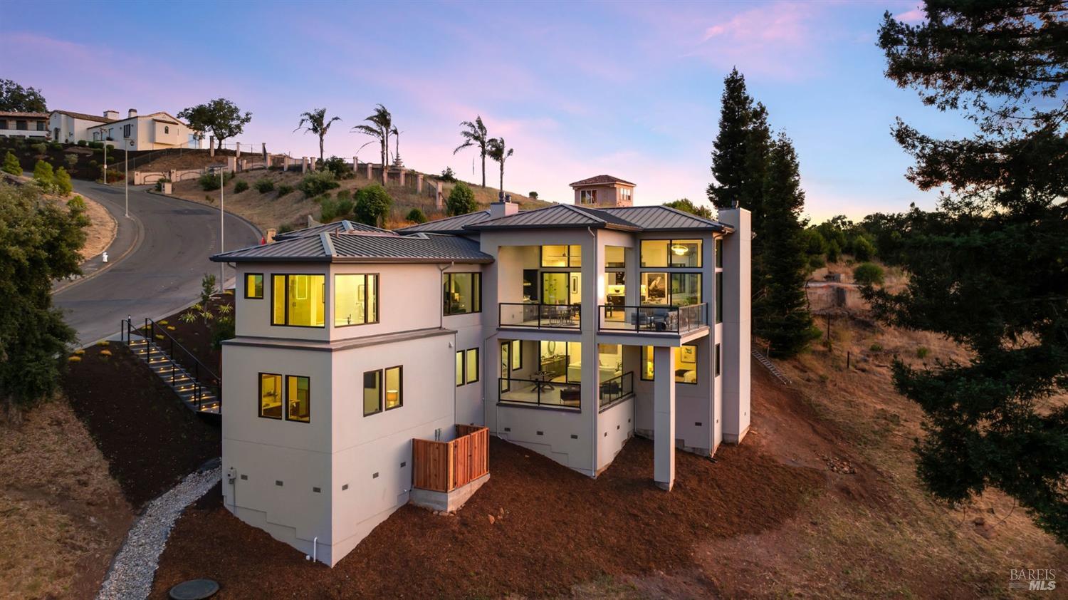 multi-story house with a metal roof, multiple balconies, and large windows, positioned on a hillside with a view of another house up the road.