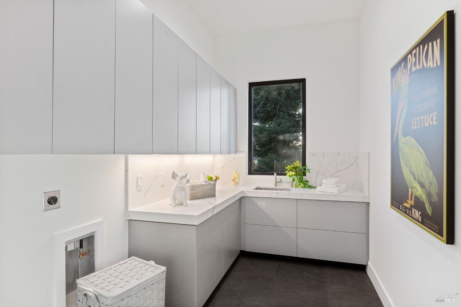 A small laundry room with modern cabinetry, a sink, and a window overlooking greenery.