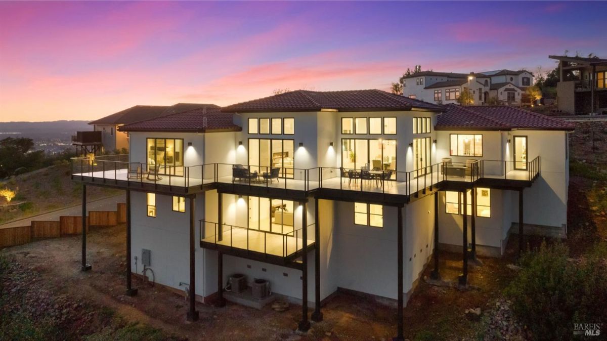 A luxurious modern home with a large balcony, elevated structure, and expansive glass windows, beautifully lit against a sunset sky.