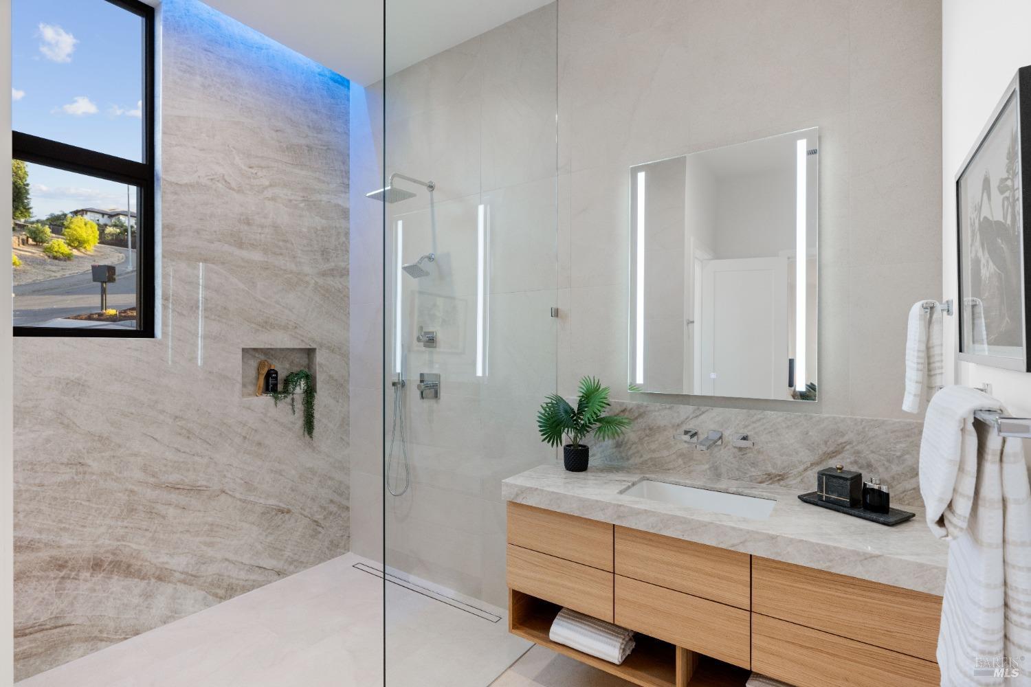 Bathroom with beige stone tile, large glass shower, and wooden vanity.