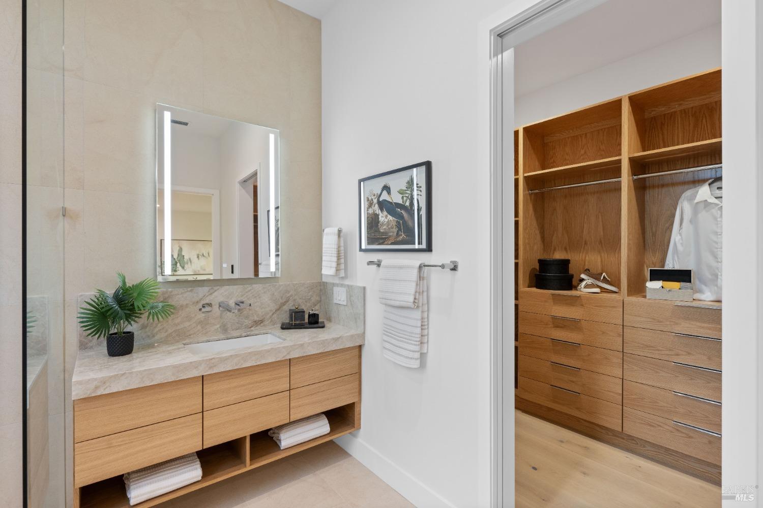 Bathroom vanity with a stone countertop, backlit mirror, and access to a walk-in closet.