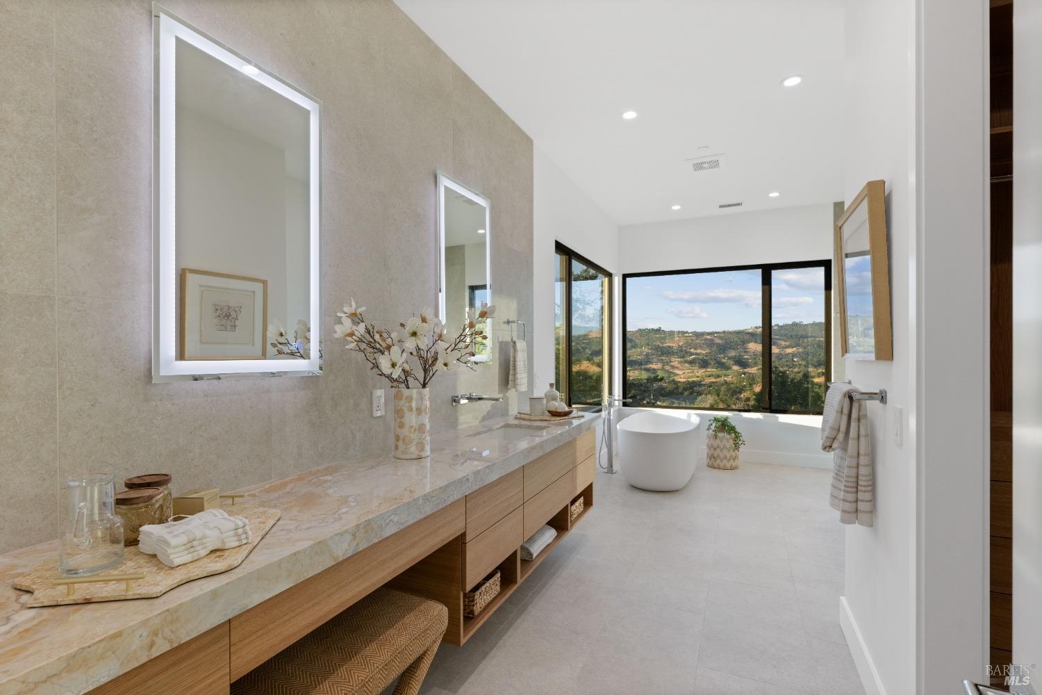 Modern bathroom with a freestanding tub, dual vanities, and large windows overlooking a scenic landscape.