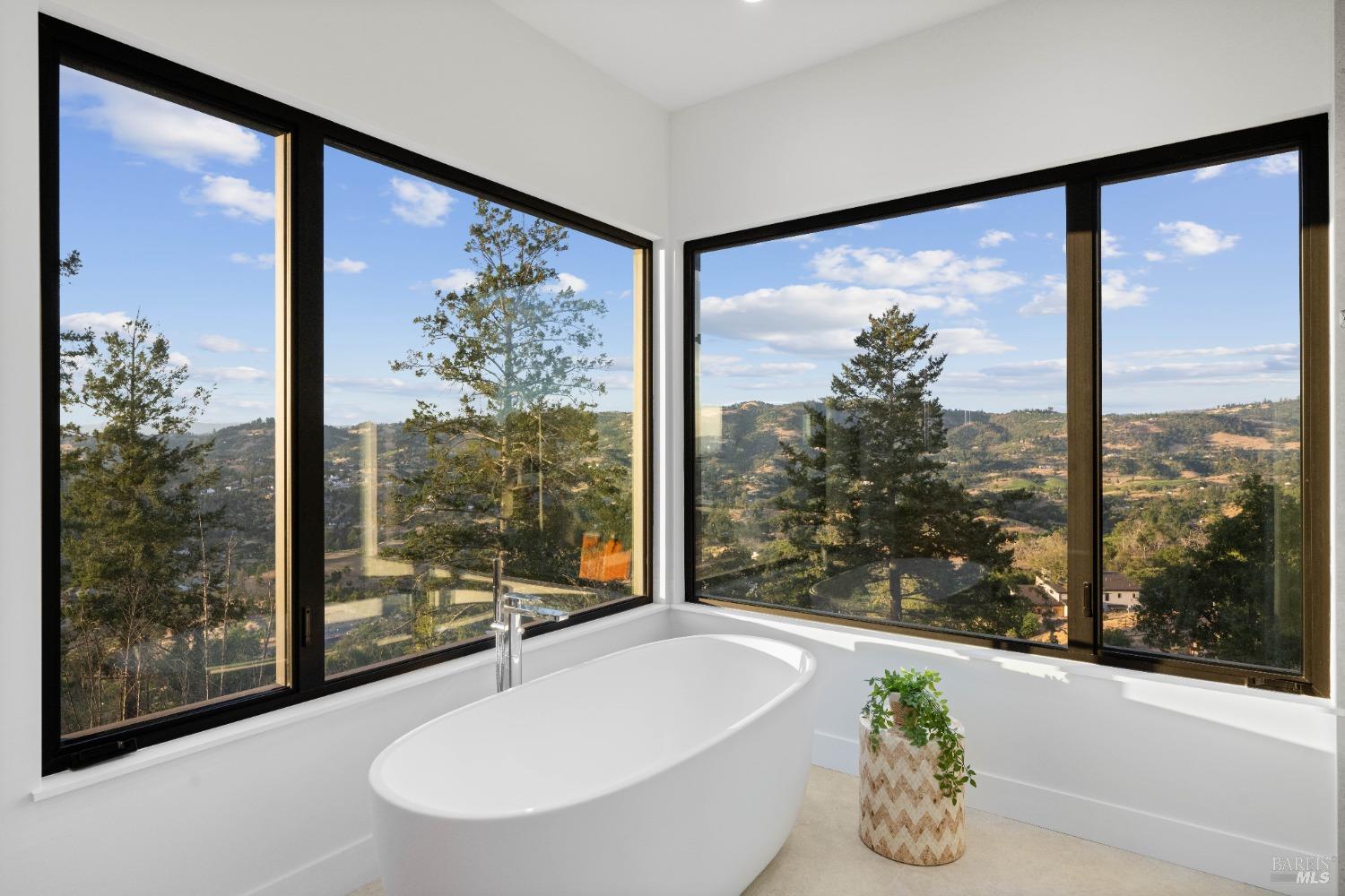 Freestanding white bathtub positioned by large corner windows overlooking a scenic view.