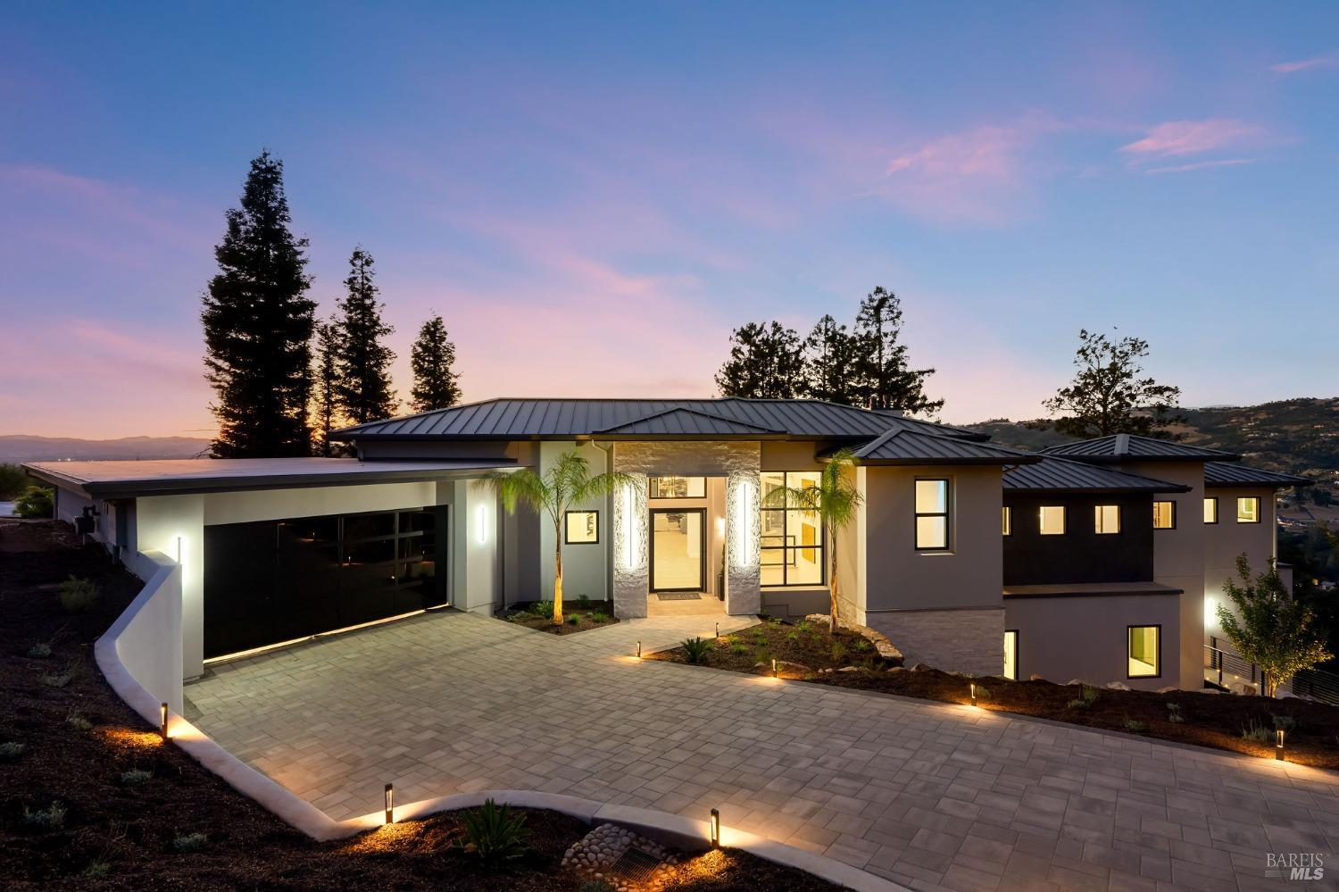 Modern house with a metal roof, a paved driveway and walkway, and illuminated landscaping at dusk.