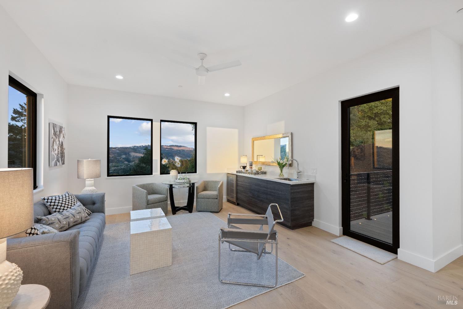 Cozy sitting area with a gray sofa, light chairs, and large windows overlooking hills.