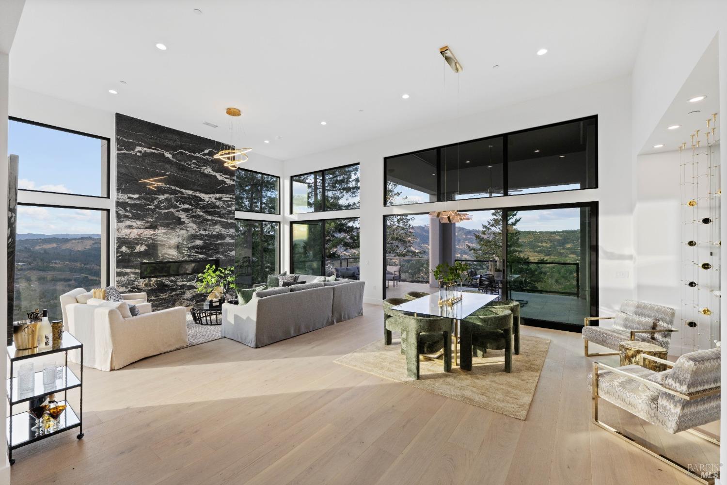 Open-concept living and dining area with a marble accent wall, high ceilings, and expansive glass windows showcasing a scenic landscape.