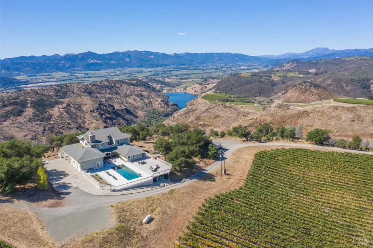 Aerial view of a house surrounded by hilly terrain and a vineyard with a nearby waterbody.