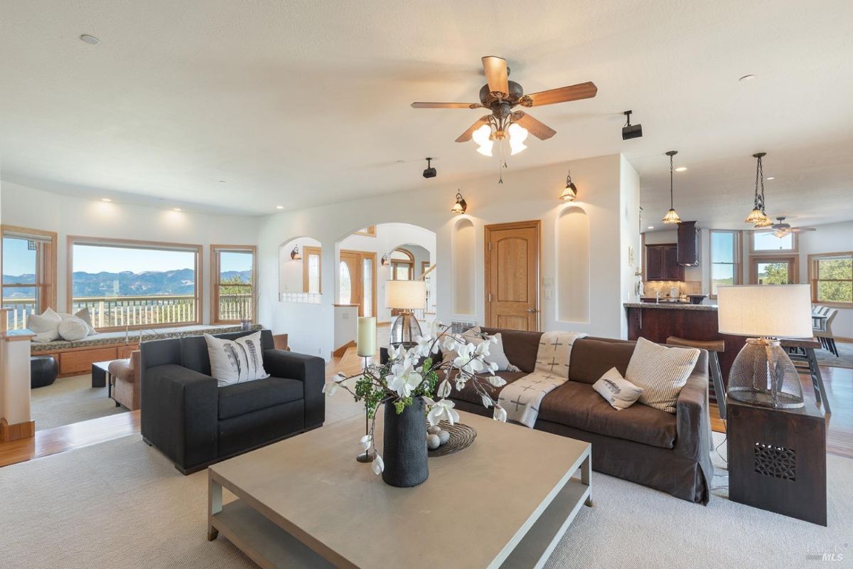 Living room with large windows, ceiling fans, and connected open spaces, including a kitchen and a seating area.