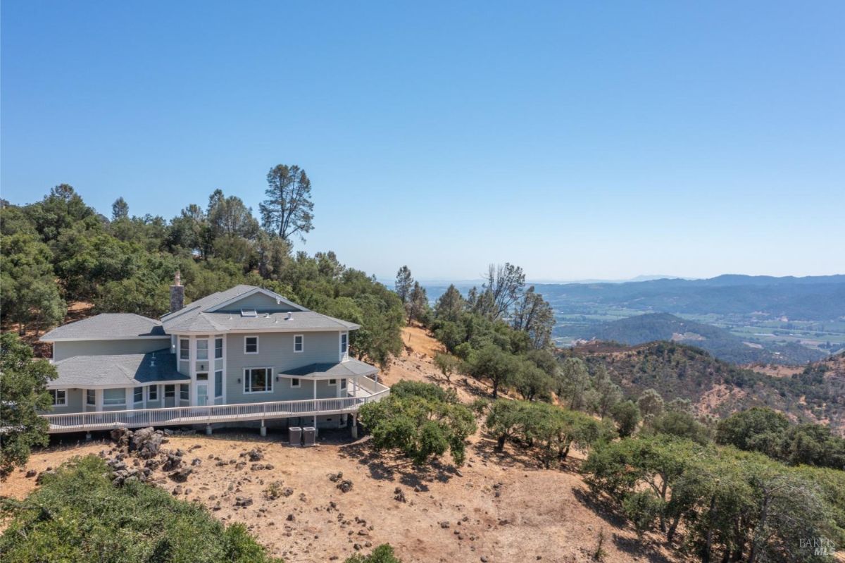 Aerial view of a hillside house with a wraparound deck and surrounding trees.